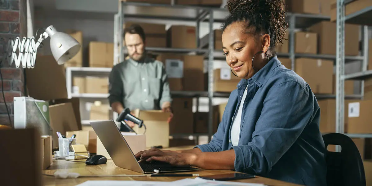Woman in warehouse sits at text and types on laptop while surrounded by boxes