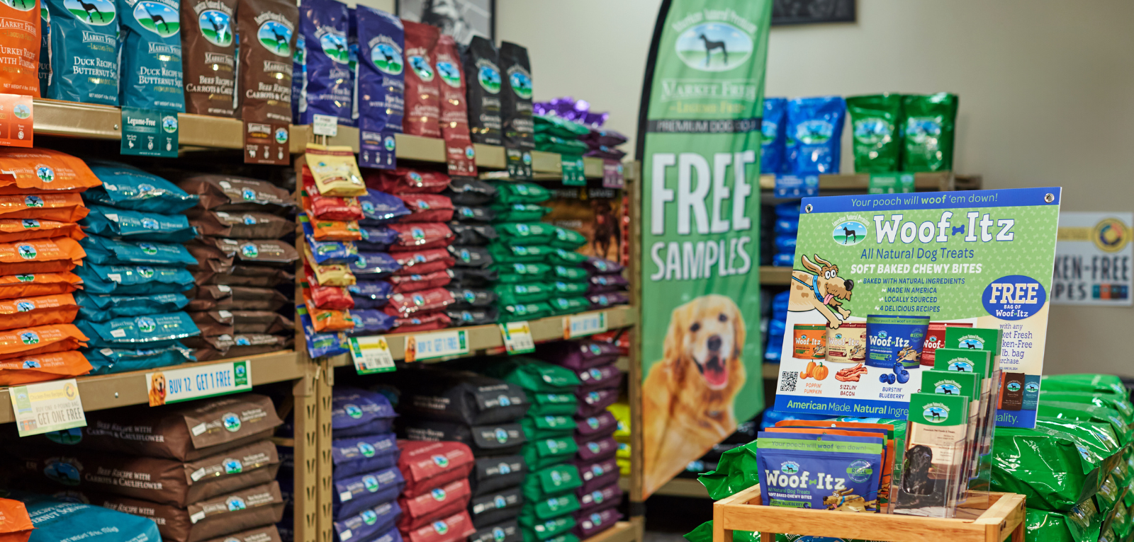 Shelves with stacks of dog food, a large flag promoting a free dog food product, a sign promoting a product, and stacks of printed marketing materials.